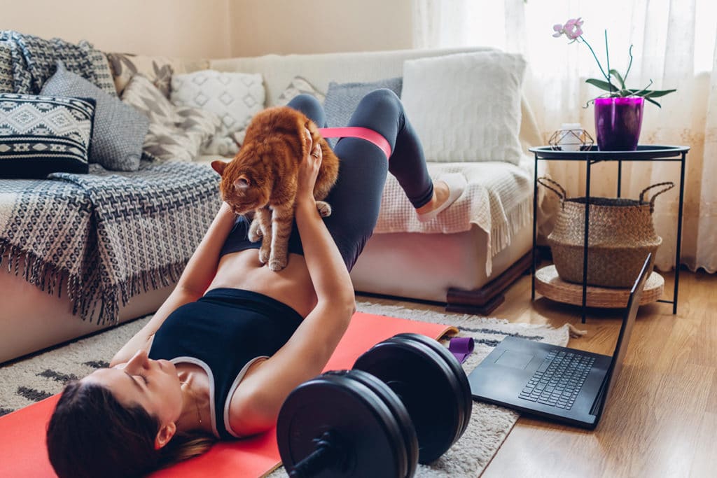 Une femme qui fait du sport avec son chat roux dans un salon. Ils sont sur un tapis de sport rouge et il y a des haltères noires et un ordinateur noir