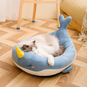 Un coussin pour chat en forme de baleine, un chat installé dedans. Le coussin est posé au sol sur un parquet en point de Hongrie, une chaise derrière et un mur blanc.