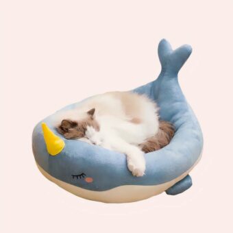 Un coussin pour chat en forme de baleine, un chat installé dedans. Le coussin est posé au sol sur un parquet en point de Hongrie, une chaise derrière et un mur blanc.