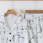 Un ensemble de pyjama chemise et chort suspendu par des cintres pendus sur un porte-manteau mural. Pyjama blanc imprimé chat. Sur un mur blanc.