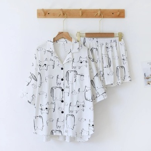 Un ensemble de pyjama chemise et chort suspendu par des cintres pendus sur un porte-manteau mural. Pyjama blanc imprimé chat. Sur un mur blanc.