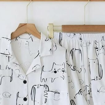 Un ensemble de pyjama chemise et chort suspendu par des cintres pendus sur un porte-manteau mural. Pyjama blanc imprimé chat. Sur un mur blanc.