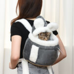 Sac à dos chat en peluche doux et chaud de couleur gris, portée par une femme debout devant un rideau blanc