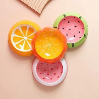 Photo de quatre gamelles pour chat, avec pour design un fruit différent chacune, nous avons pastèque, orange, fraise et citrouille, la citrouille est posée au centre et au-dessus des trois autres, le tout posé sur une table de couleur saumon