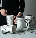Une femme debout, habillée en noir, tiens dans sa main un des quatre mugs de grande taille, avec une cuillère à l'intérieur, les mugs sont noir et blanc, deux ont un couvercle noir, ayant pour poignée une forme de queue de chat, chacun ont un dessin différent d'un noir gris et blanc, ils sont posés sur une table, l'un des mug est posé couché avec une cuillère à l'intérieur, la table est grise, on peut y voir des grains de café et un morceau d'écharpe grise dessus.