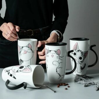 Une femme debout, habillée en noir, tiens dans sa main un des quatre mugs de grande taille, avec une cuillère à l'intérieur, les mugs sont noir et blanc, deux ont un couvercle noir, ayant pour poignée une forme de queue de chat, chacun ont un dessin différent d'un noir gris et blanc, ils sont posés sur une table, l'un des mug est posé couché avec une cuillère à l'intérieur, la table est grise, on peut y voir des grains de café et un morceau d'écharpe grise dessus.