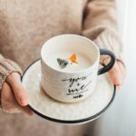 Une femme habillée d'un pull beige, tient dans ses mains, un mug posé sur sa soucoupe, le tout en céramique, le mug et la soucoupe sont blancs avec un liseré noir. La tasse à une inscription écrite "you + me = petit dessin de poisson" il y a du lait dedans et il ressort un petit chat en relief roux blanc et gris