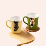 Il y a posé sur une table, un plateau avec un mug jaune qui a pour design un chat noir qui grimpe dessus, à côté, nous avons sur un dessous de verre un mug vert ayant un chat blanc, noir et roux qui grimpe sur celui-ci.