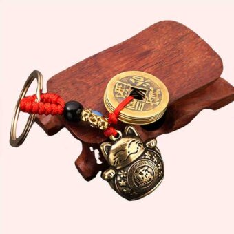 Porte-clé doré en forme de chat, du type Bouddha, ayant des inscriptions chinoises sur le ventre, il lève ses deux pattes, il a un sourire est les yeux fermés, il y a aussi un anneau doré accroché avec des écritures chinoises, il est attaché par un cordon rouge, décoré d'une petite perle noire et un petit ornement doré et bleu, il est posé sur un petit morceau de bois, le tout sur un fond noir