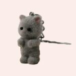 Petit porte-clé chat debout, gris, tout poilu, qui parait tout doux, est posé sur une table en bois, l'anneau qui est relié par une petite chaine, est posé sur un plat blanc en verre