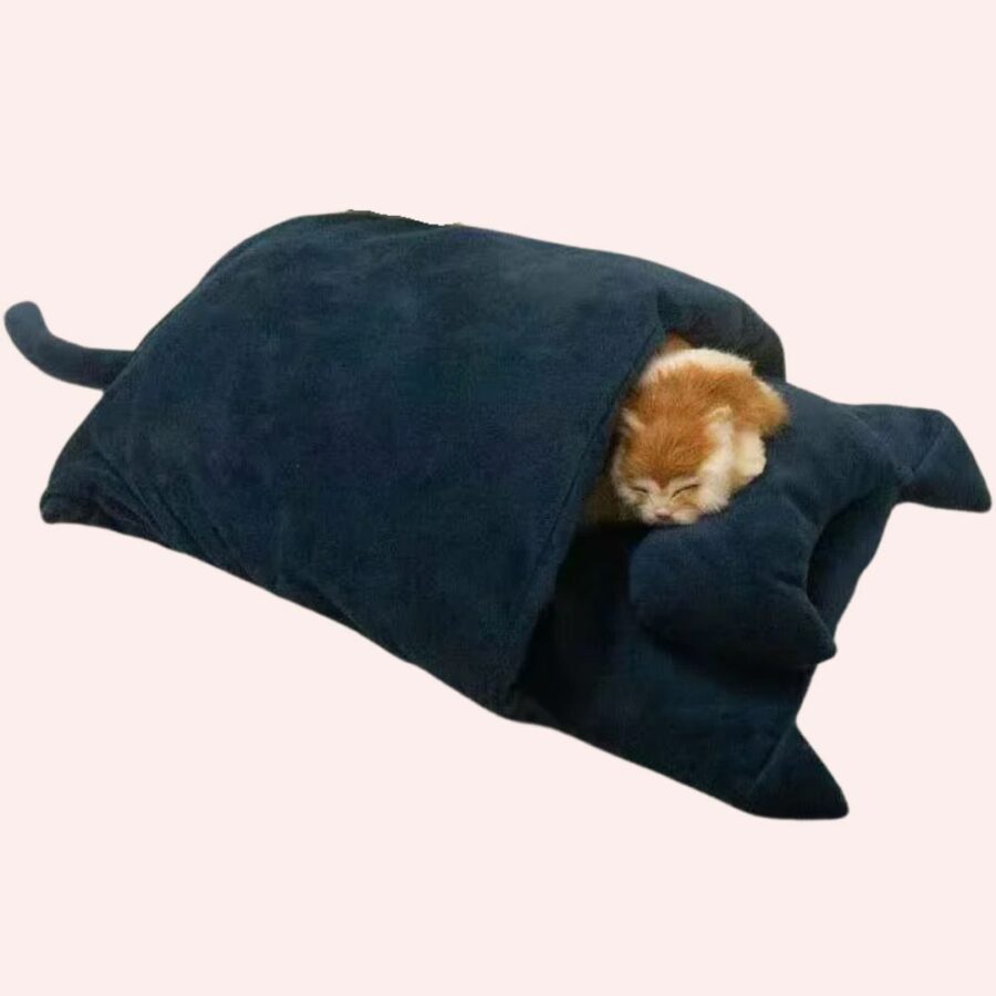 Sac de couchage bleu velours forme chat avec un chat roux qui dort dedans sur fond rose.